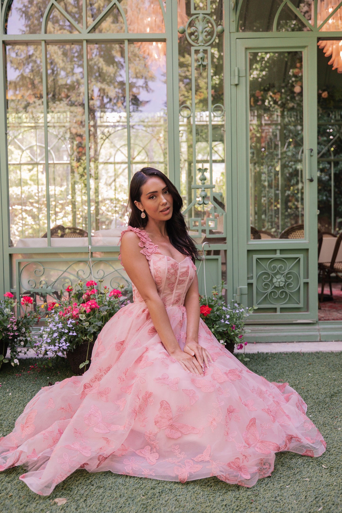 The image shows a woman in a pink dress sitting on the ground in front of a glass door, surrounded by plants with flowers and trees in the background.