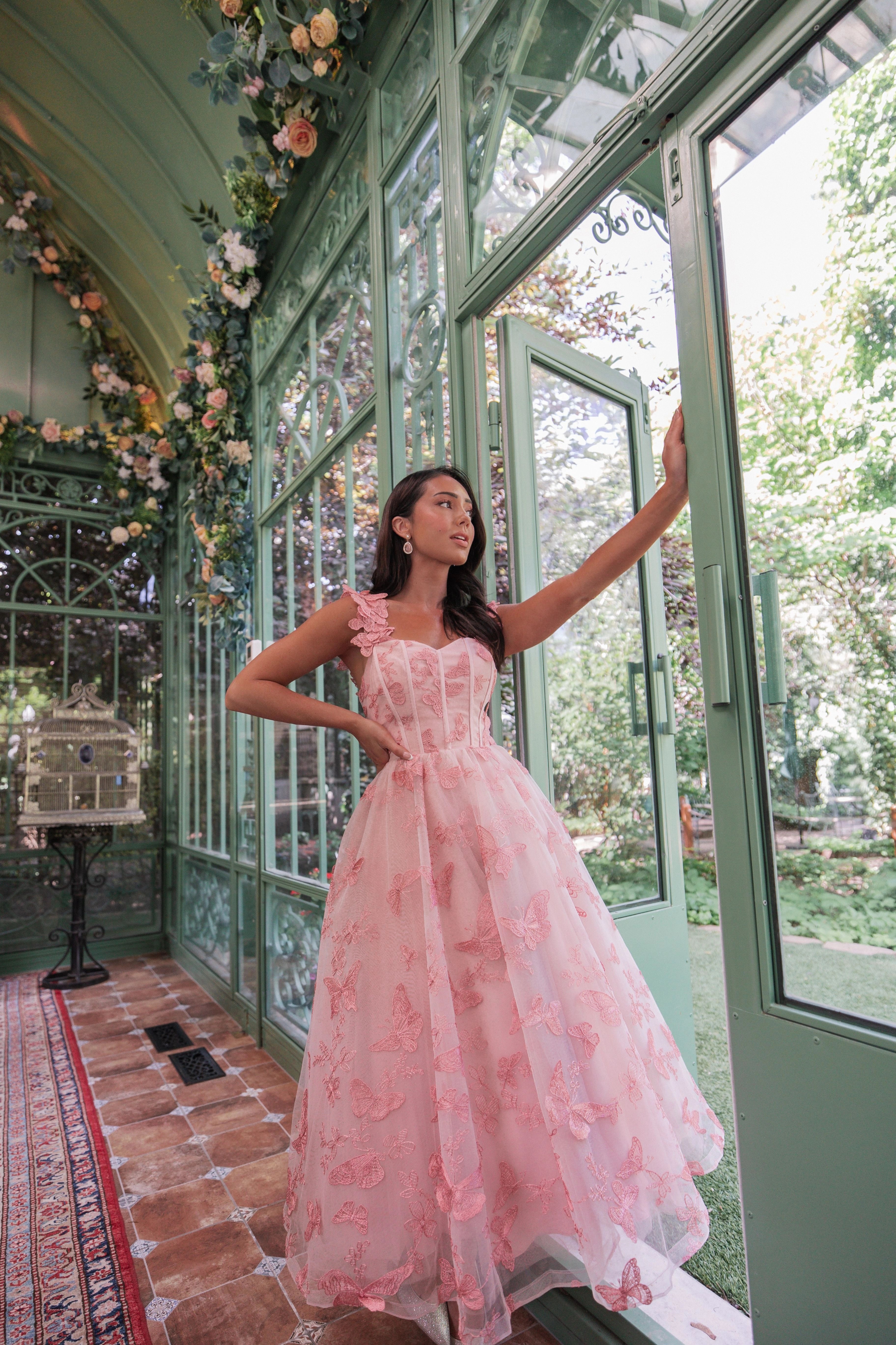 The image shows a woman in a beautiful pink wedding dress standing in front of a glass door, surrounded by a carpeted floor, a table, a chair, and a few flowers and leaves attached to the wall.