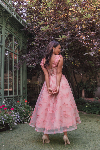 The image shows a woman in a pink dress standing in front of a green house surrounded by plants with flowers and trees. She is wearing a dress that is both pink and has a flower in her hand.