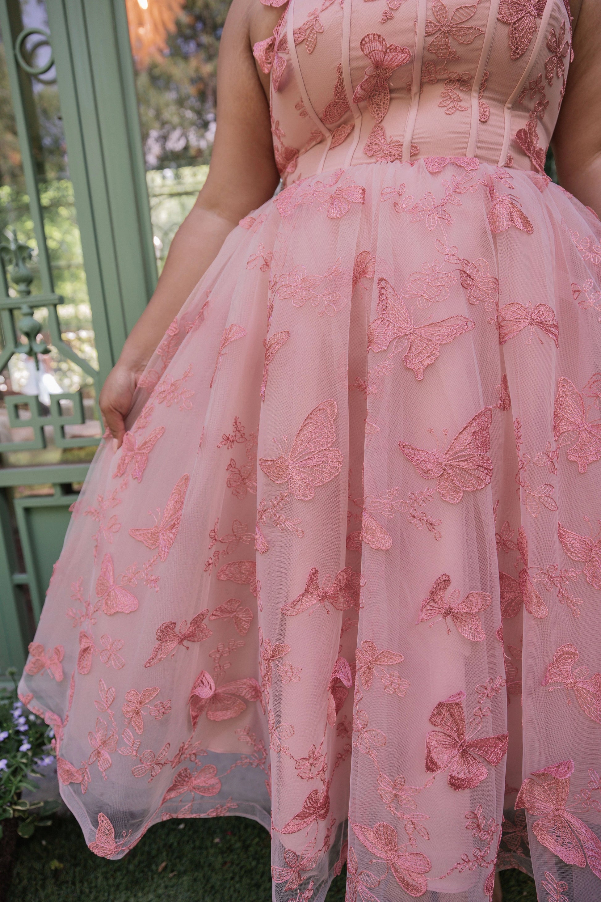 The image shows a woman in a pink dress standing in front of a gate, with a few plants in the background. She is wearing a pink butterfly dress, giving the image a whimsical and dreamy feel.