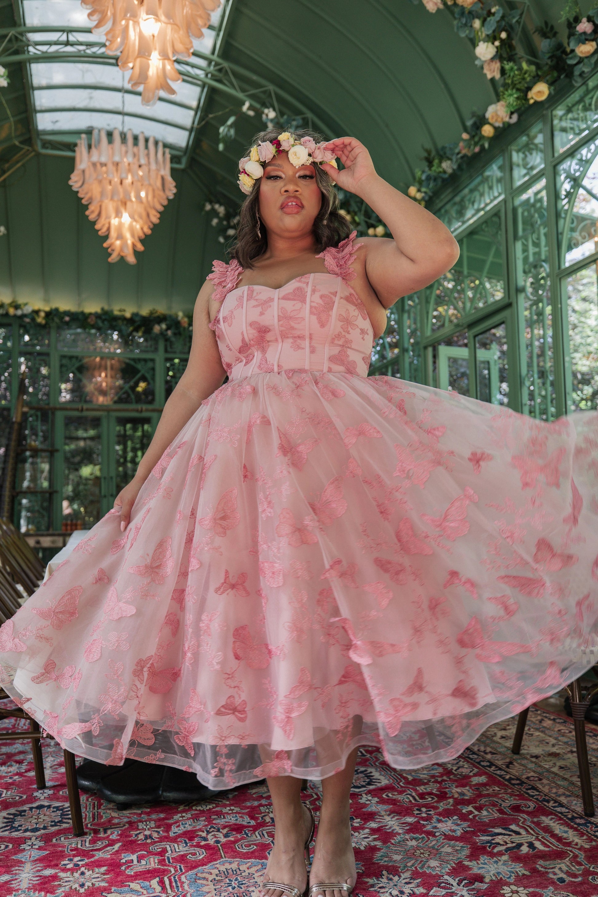 The image shows a woman in a pink dress standing on a red carpet, surrounded by chairs, flowers, and chandeliers. She is wearing a crown on her head, and the background is slightly blurred.