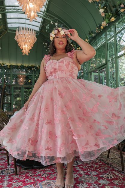 The image shows a woman in a pink dress standing on a red carpet, surrounded by chairs, flowers, and chandeliers. She is wearing a crown on her head, and the background is slightly blurred.