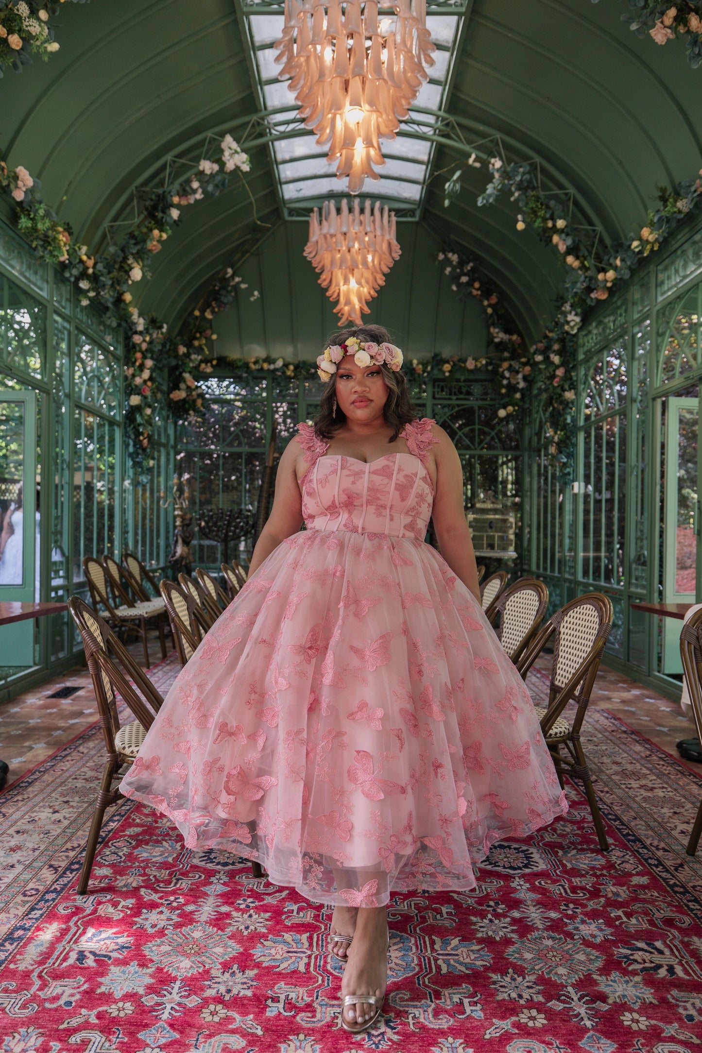 The image shows a woman in a pink dress standing on a red carpet in a room with chairs, a chandelier, and a glass wall in the background.