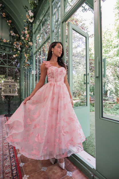 The image shows a woman in a pink dress standing in front of a glass door, surrounded by a carpeted floor, a table, and a few flowers and leaves. Through the glass door we can see trees and plants in