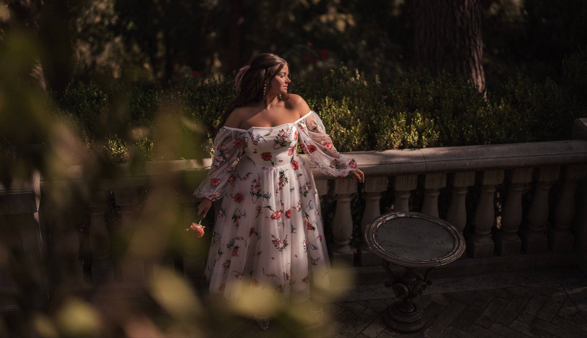 Woman in a floral off-shoulder dress standing in a garden setting.