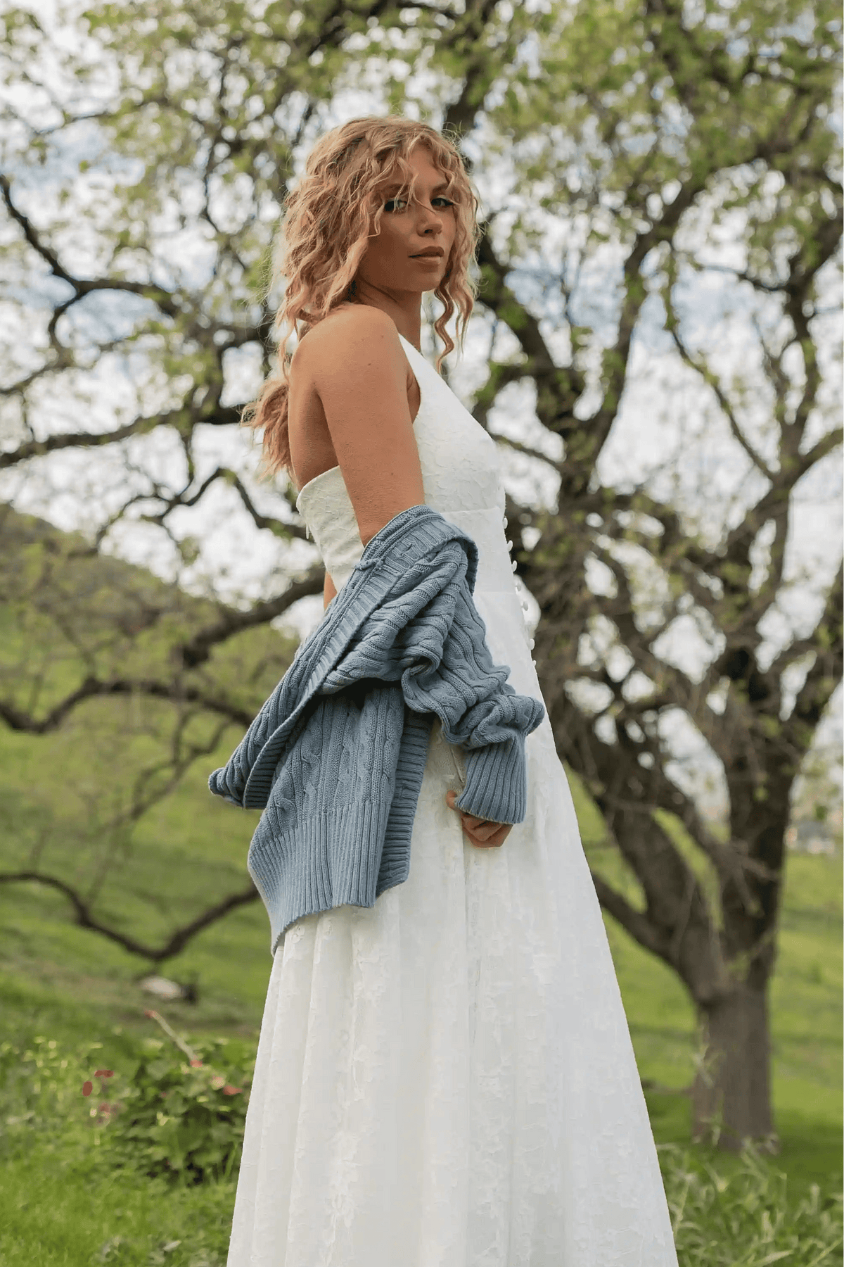Woman in a white dress with a blue cardigan standing in a natural setting with trees and greenery.