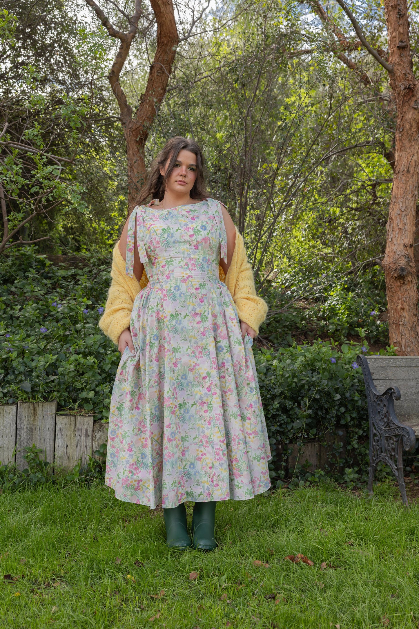 Woman in floral dress and yellow cardigan standing in grassy area with trees and wooden fence behind her.