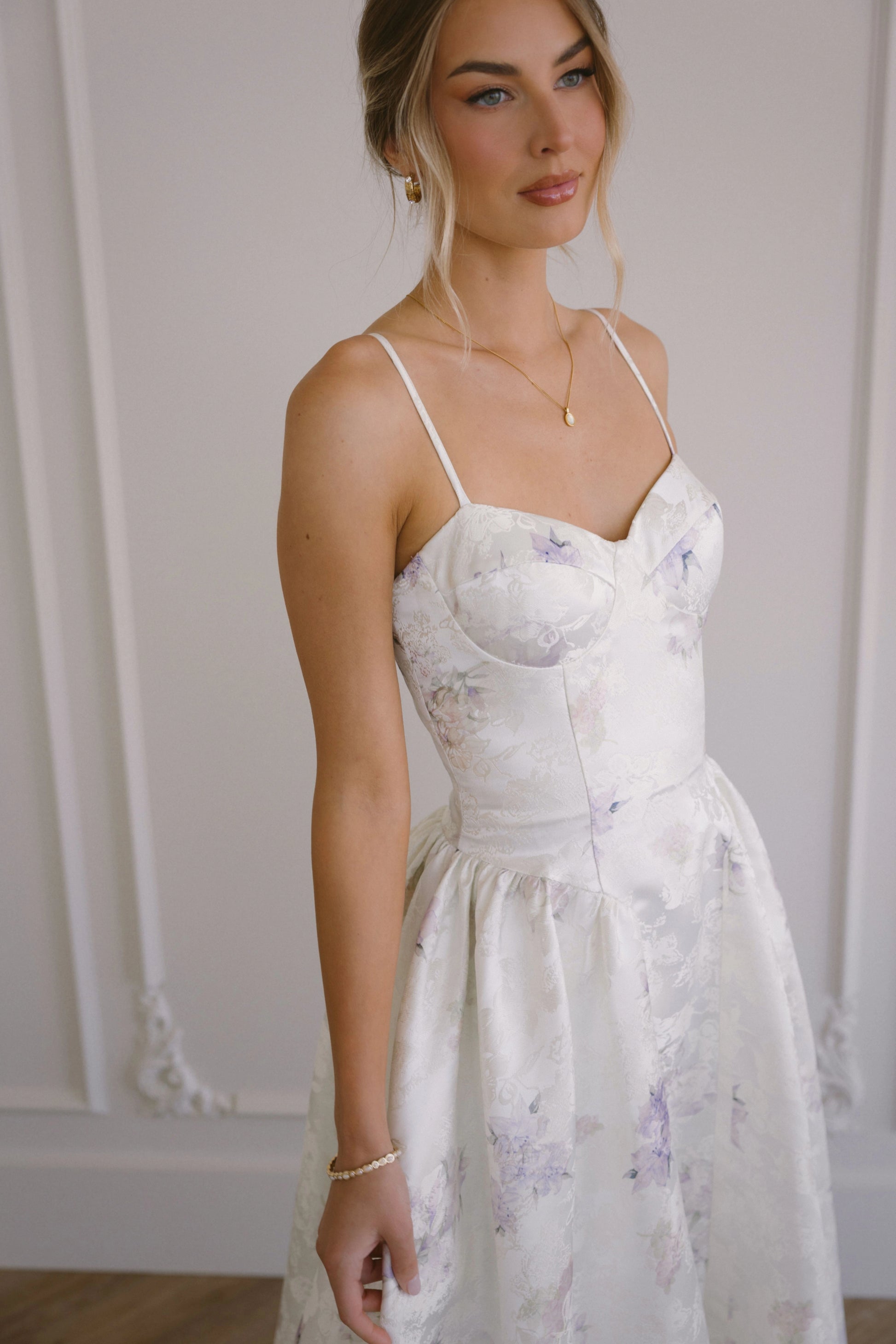 Woman in a white floral dress with thin straps and sweetheart neckline standing in front of a white wall.