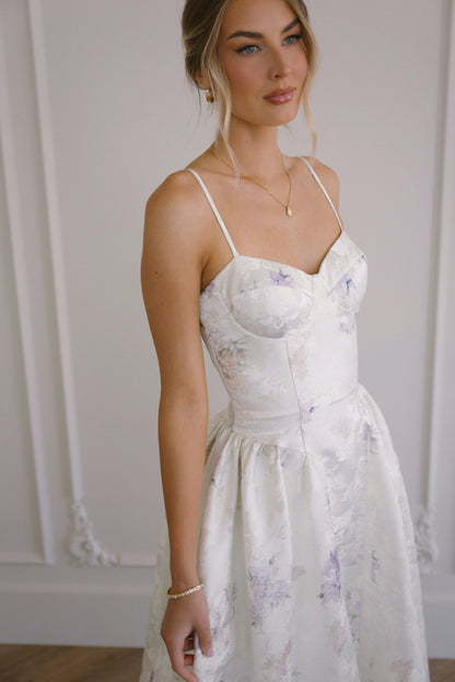 Woman in a white floral dress with thin straps and sweetheart neckline standing in front of a white wall.