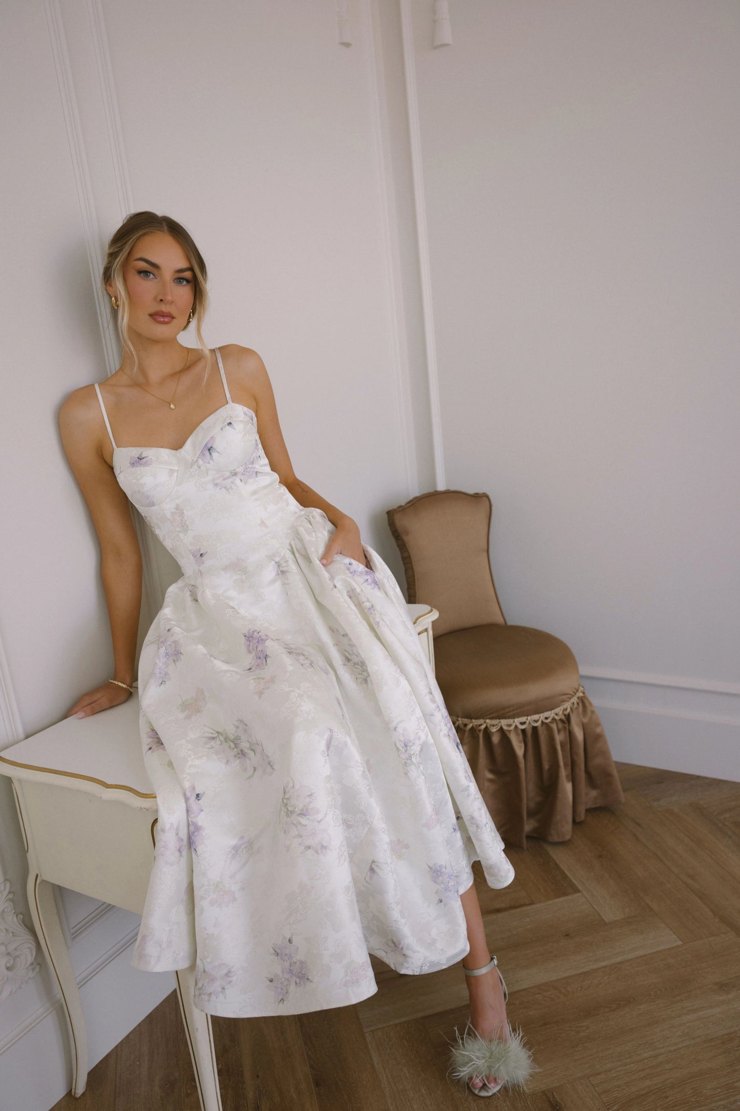 Woman in white floral dress with spaghetti straps sits on vanity beside brown chair in white room.