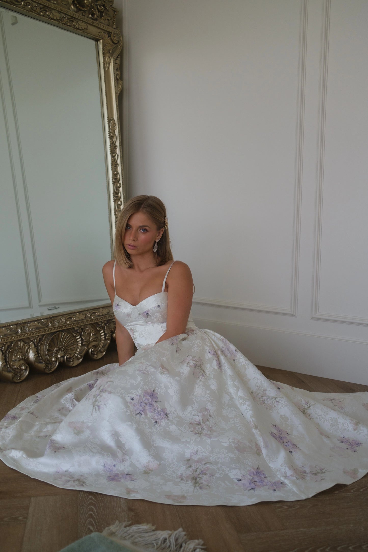 Woman in white floral wedding dress with spaghetti straps sits on wooden floor beside large ornate gold-framed mirror.
