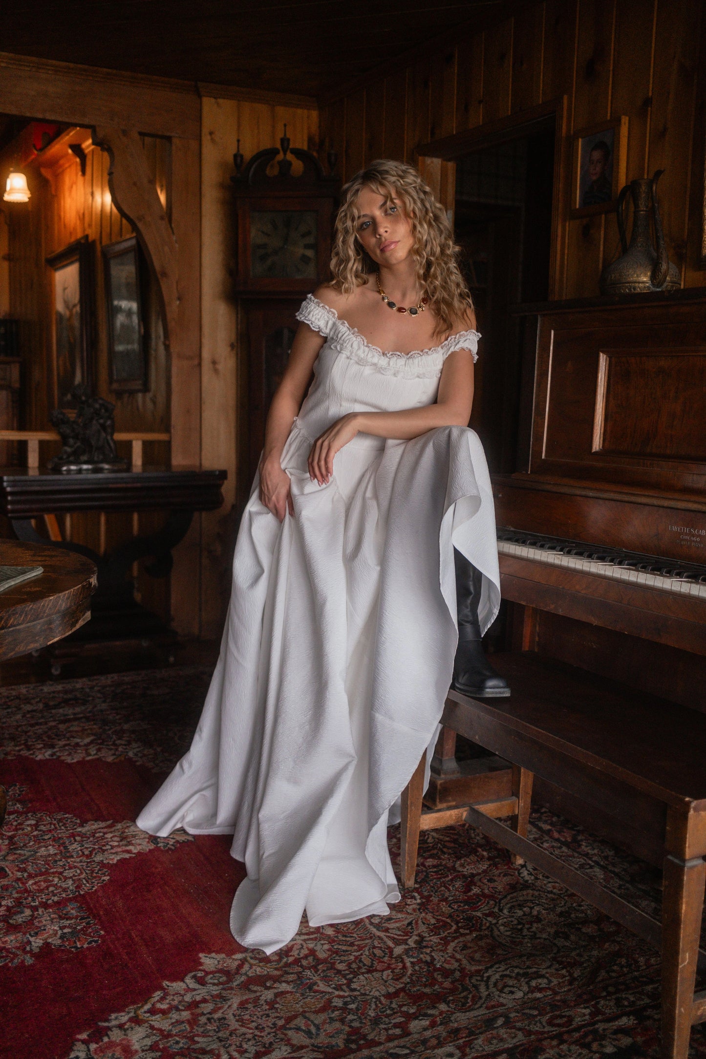 Woman in white off-the-shoulder dress sits on piano bench beside dark wood piano in a wood-paneled room with ornate rug.