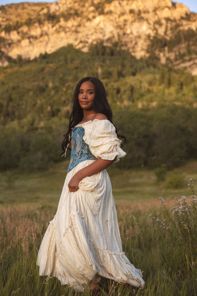 The image shows a woman in a white dress standing in a field of tall grass, with a hill in the background and a clear blue sky above.