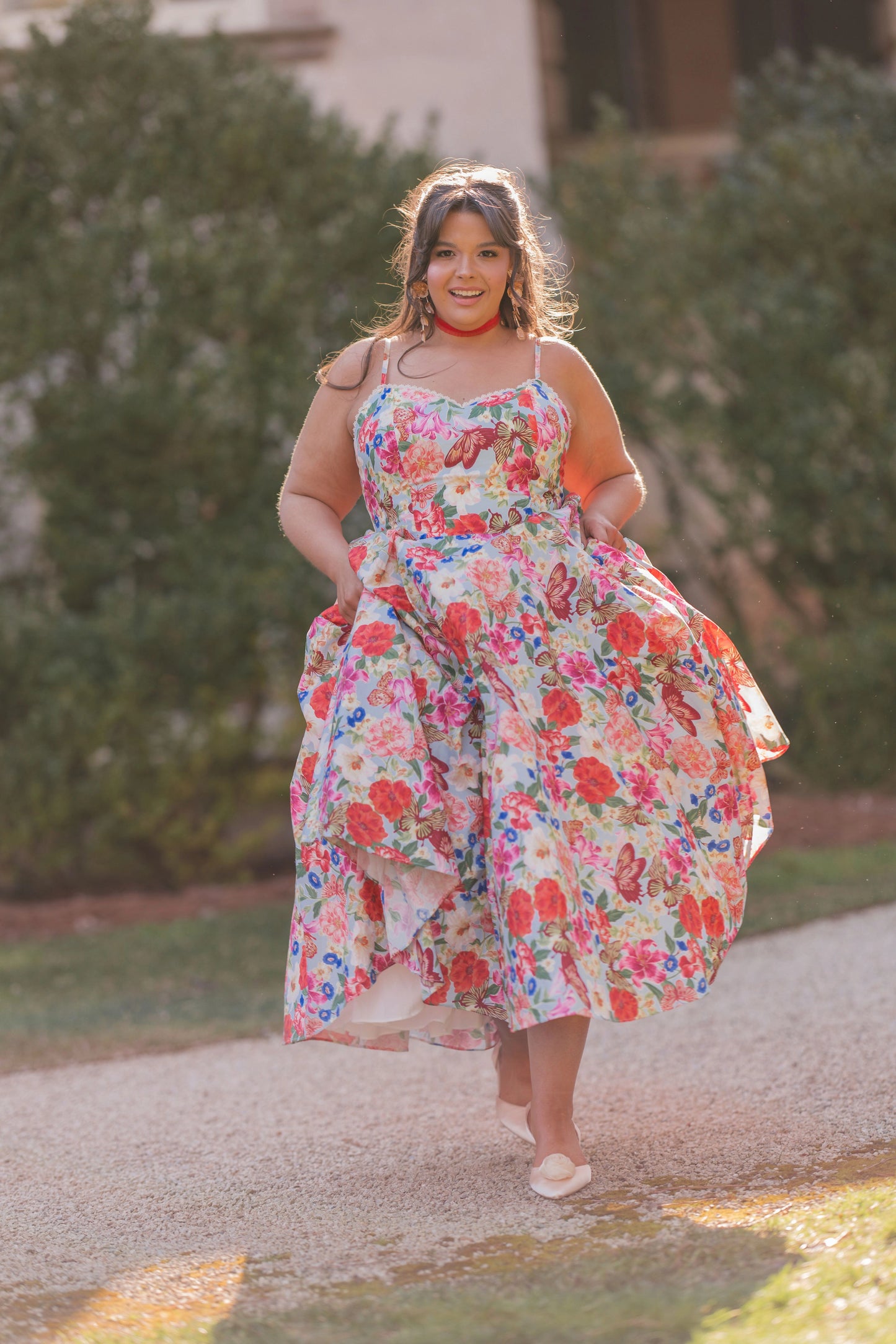 The image shows a woman in a floral dress walking down a path surrounded by trees and a building in the background. She is wearing a pair of footwear, suggesting she is ready to take on the world.