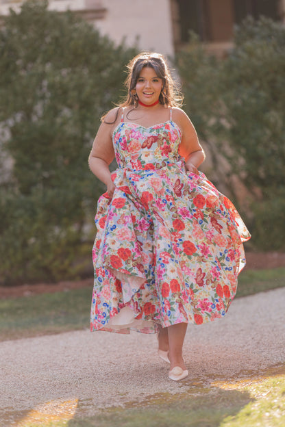 The image shows a woman in a floral dress walking down a path surrounded by trees and a building in the background. She is wearing a pair of footwear, suggesting she is ready to take on the world.