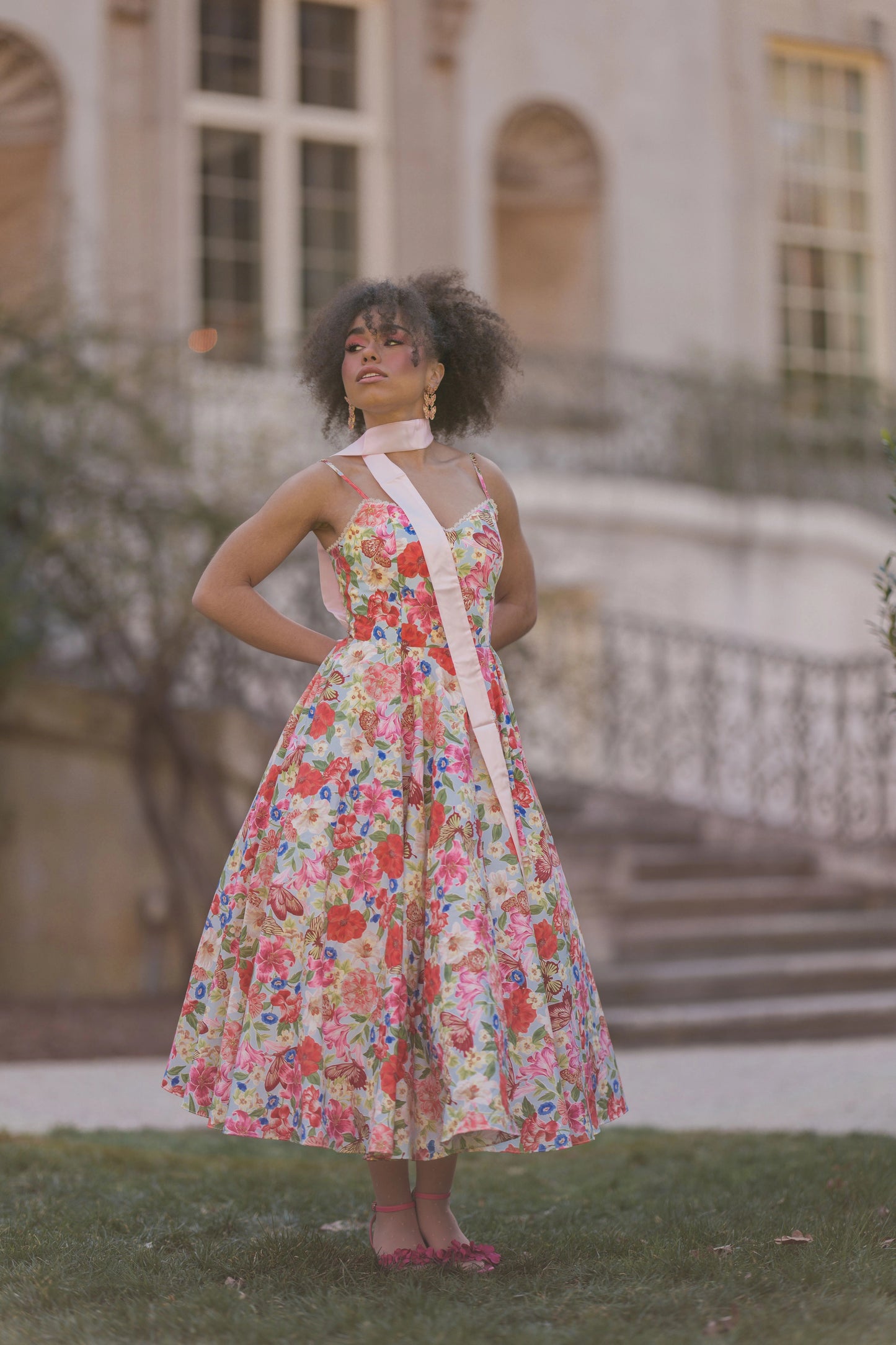 The image shows a woman in a colorful dress standing in front of a building with a tree in the background. The dress is a vibrant mix of colors, including blues, greens, yellows, and pinks, and is