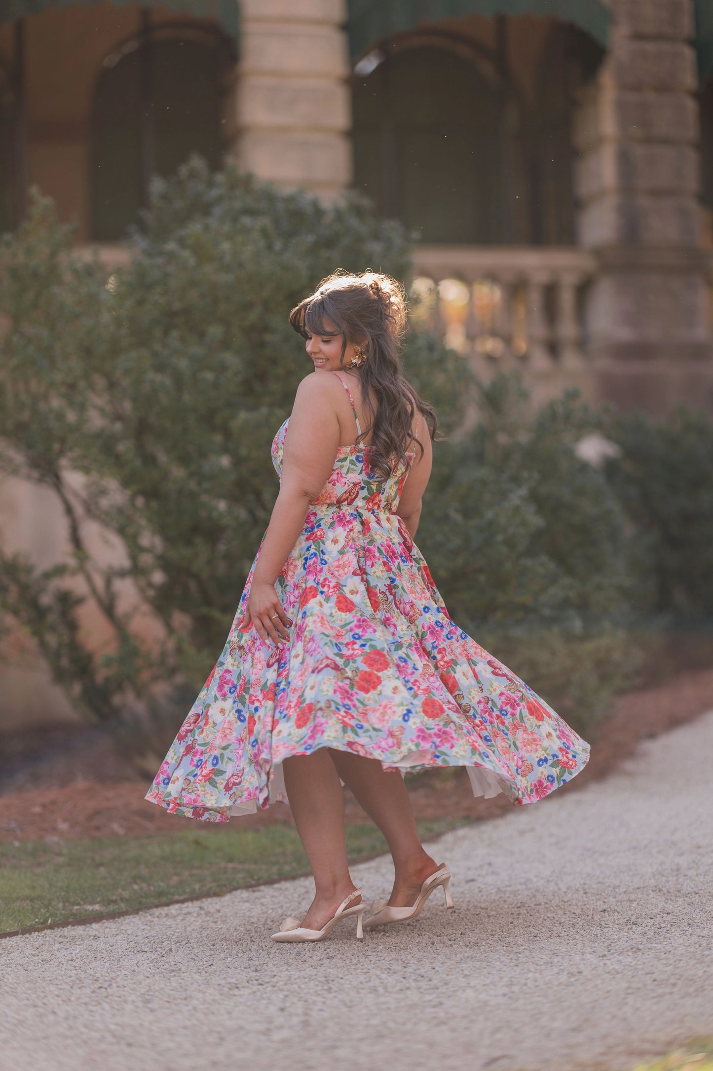 The image shows two women walking down a sidewalk in front of a building, surrounded by trees. One of the women is wearing a pair of high heels and a necklace.