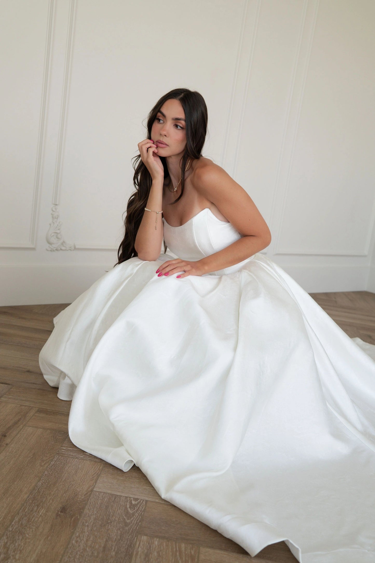 The image shows a woman in a white wedding dress sitting on the floor in front of a wall. She is wearing a white dress with intricate details and a long train. Her hair is styled in an elegant updo